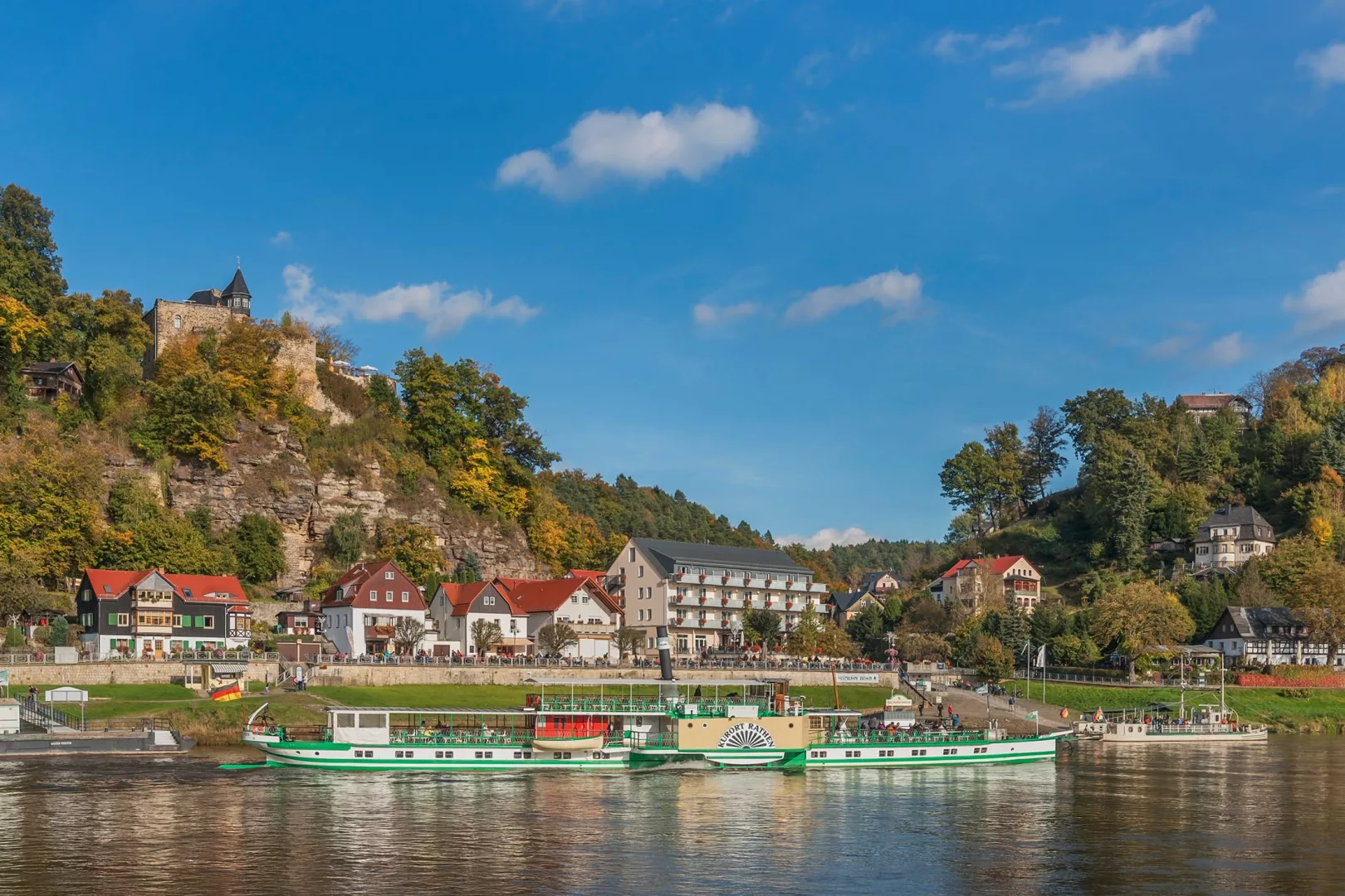 Bild von dem Dorf Rathen mit der Elbe, den Schiffen, dem Kurort und der Burg im Hintergrund, hinter der die Felsenbühne liegt