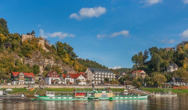 Bild von dem Dorf Rathen mit der Elbe, den Schiffen, dem Kurort und der Burg im Hintergrund, hinter der die Felsenbühne liegt