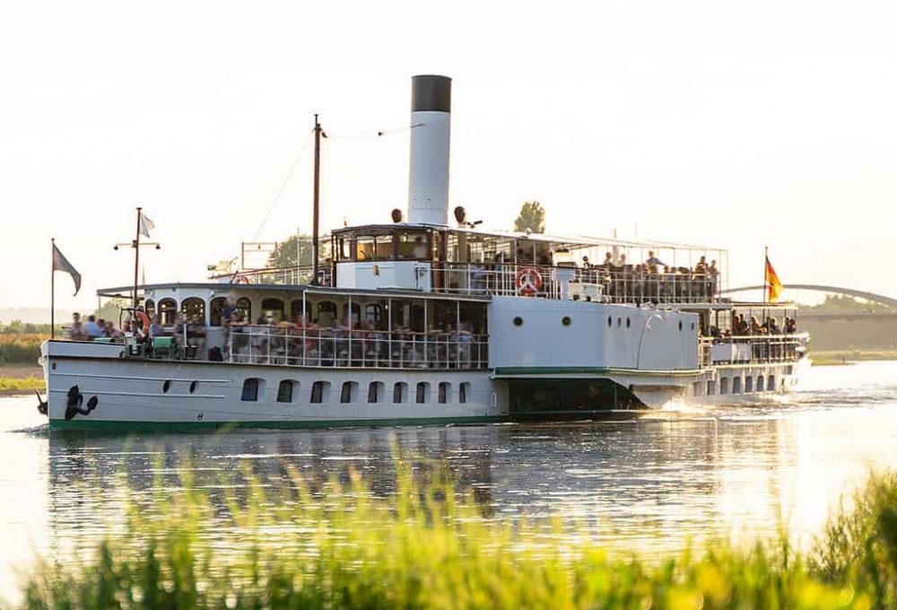 Large retro tourist steamer with a pipe is full of people and sails along the calm Elbe river next to a green wild coast on a sunny day at sunset in the city of Dresden. Travel concept